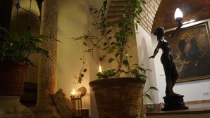 A statue of a woman holding a torch, a potted plant, and a lit candle on a stone bench in a room with a brick wall and a painting.