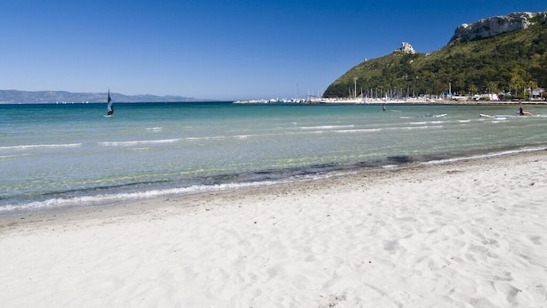 En strand med klart vatten, en sandstrand och en kulle med vegetation i bakgrunden.