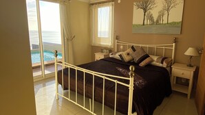 Quarto com cama, mesa de cabeceira, luminária, pintura, janela com cortinas e vista para o mar.