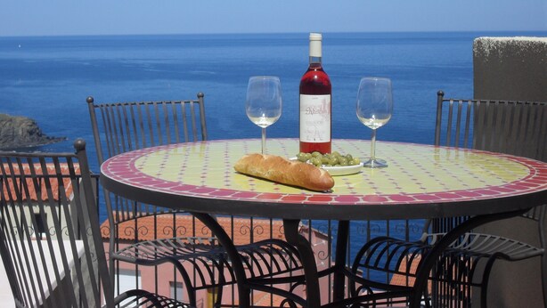 Mesa com uma garrafa de vinho, duas taças e pão, com o cenário da costa ao fundo.