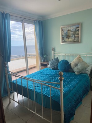 A bedroom with a bed, a painting, and a view of the sea.