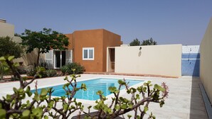 Ein Schwimmbecken mit gefliestem Deck, eine kleine Grünfläche mit Pflanzen und ein Gebäude mit beiger Fassade und weißen Fensterläden.