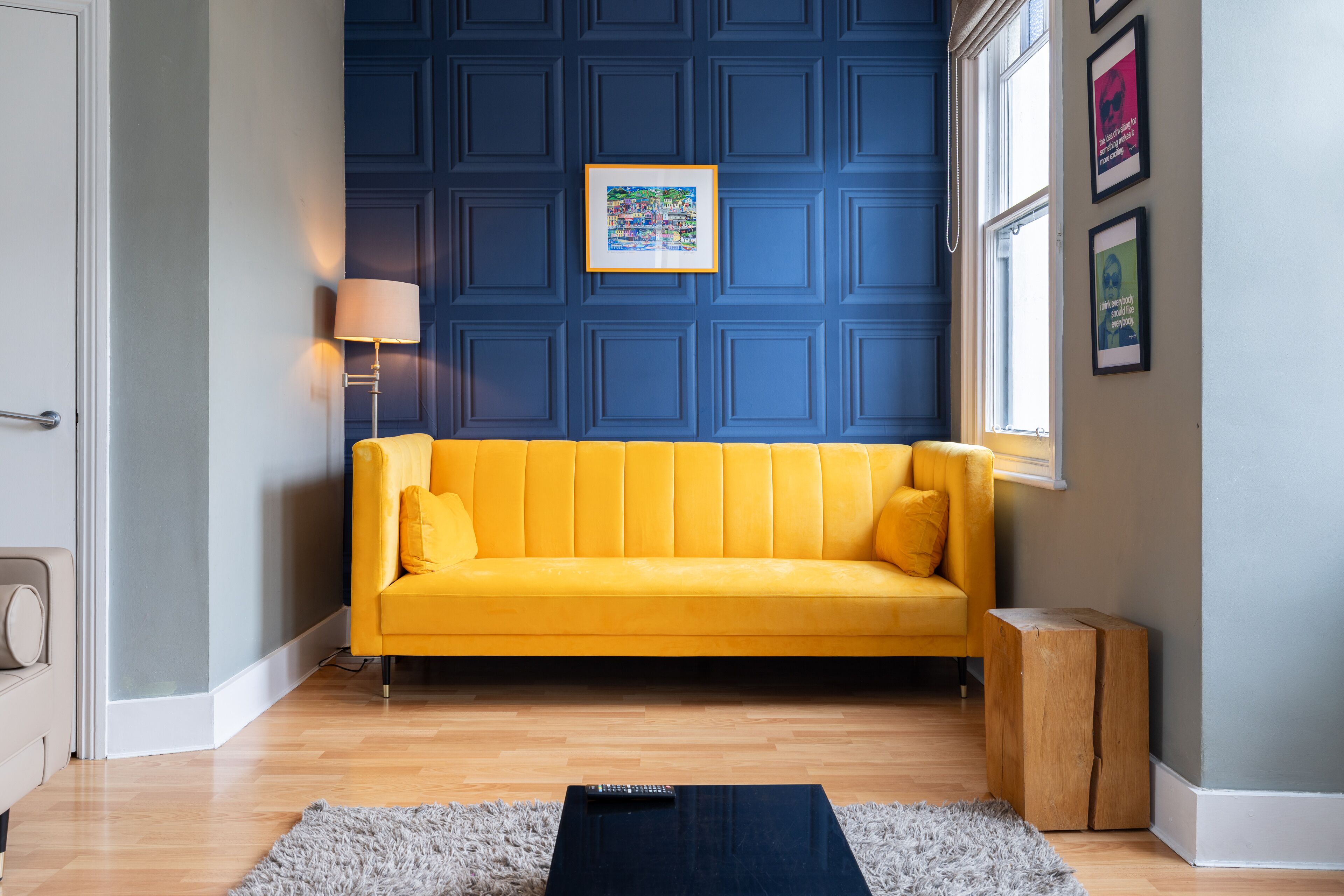Une pièce avec un canapé jaune, une table d’appoint en bois, un tableau encadré au mur et une fenêtre.