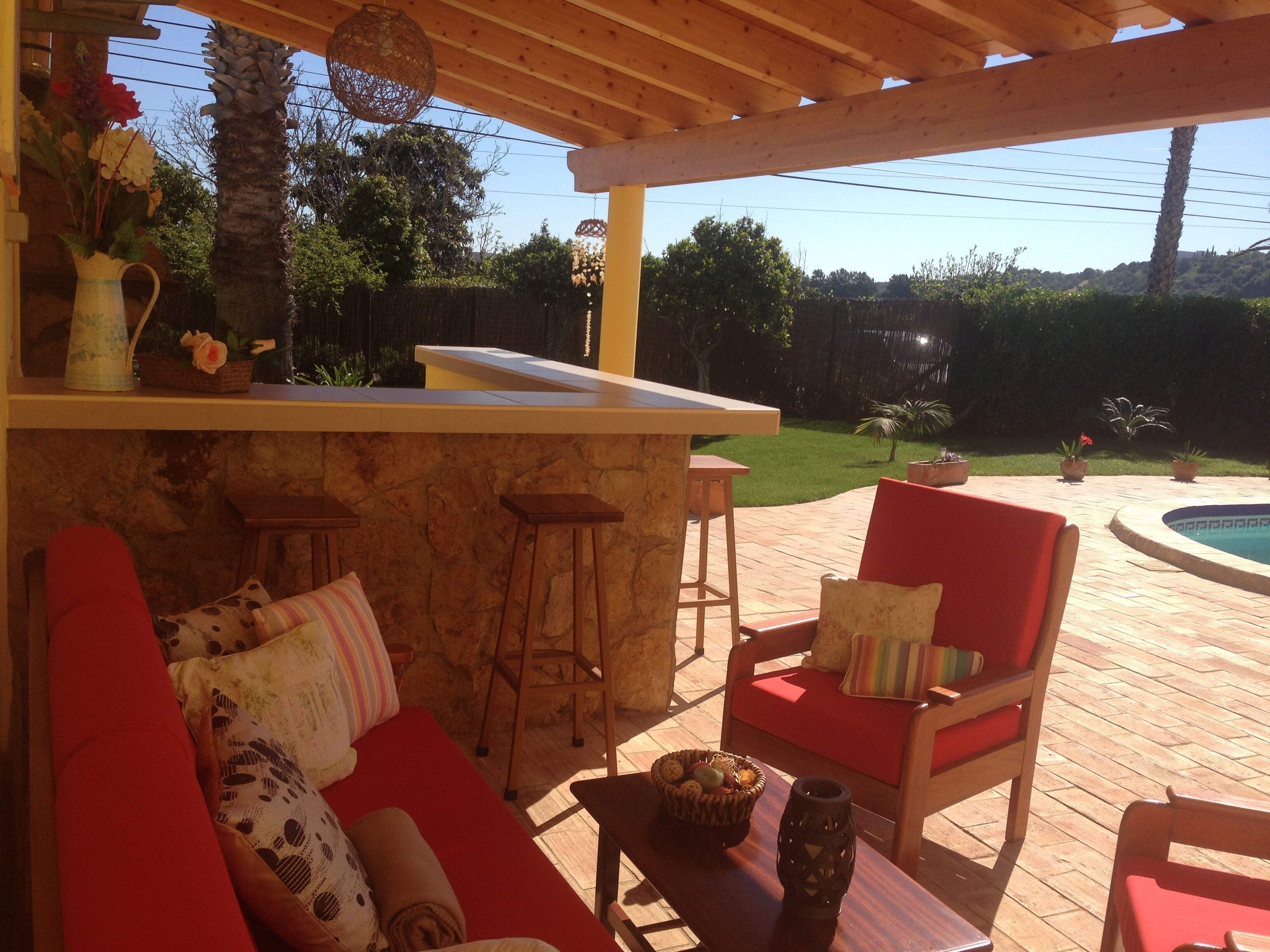 An outdoor patio with a wooden pergola, a pool, and a seating area with red cushions and a stone wall.