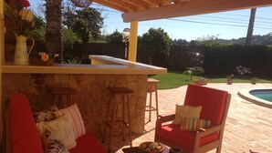 Un patio exterior con una pérgola de madera, una piscina y una zona de estar con cojines rojos y un muro de piedra.