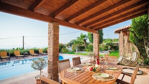A wooden outdoor dining area with a pool view, stone pillars, and a hanging chair. - Luxurious villa, idyllic view, 5km from town & beach, total privacy, large pool (Rethimno)