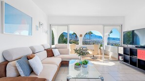 A modern living room with a sofa, coffee table, and a TV. There are large windows with a view of palm trees and mountains. - Private Garden & pool, Sea View, near restaurant, free Wi-fi, 5,5 km from Beach (La Matanza de Acentejo)