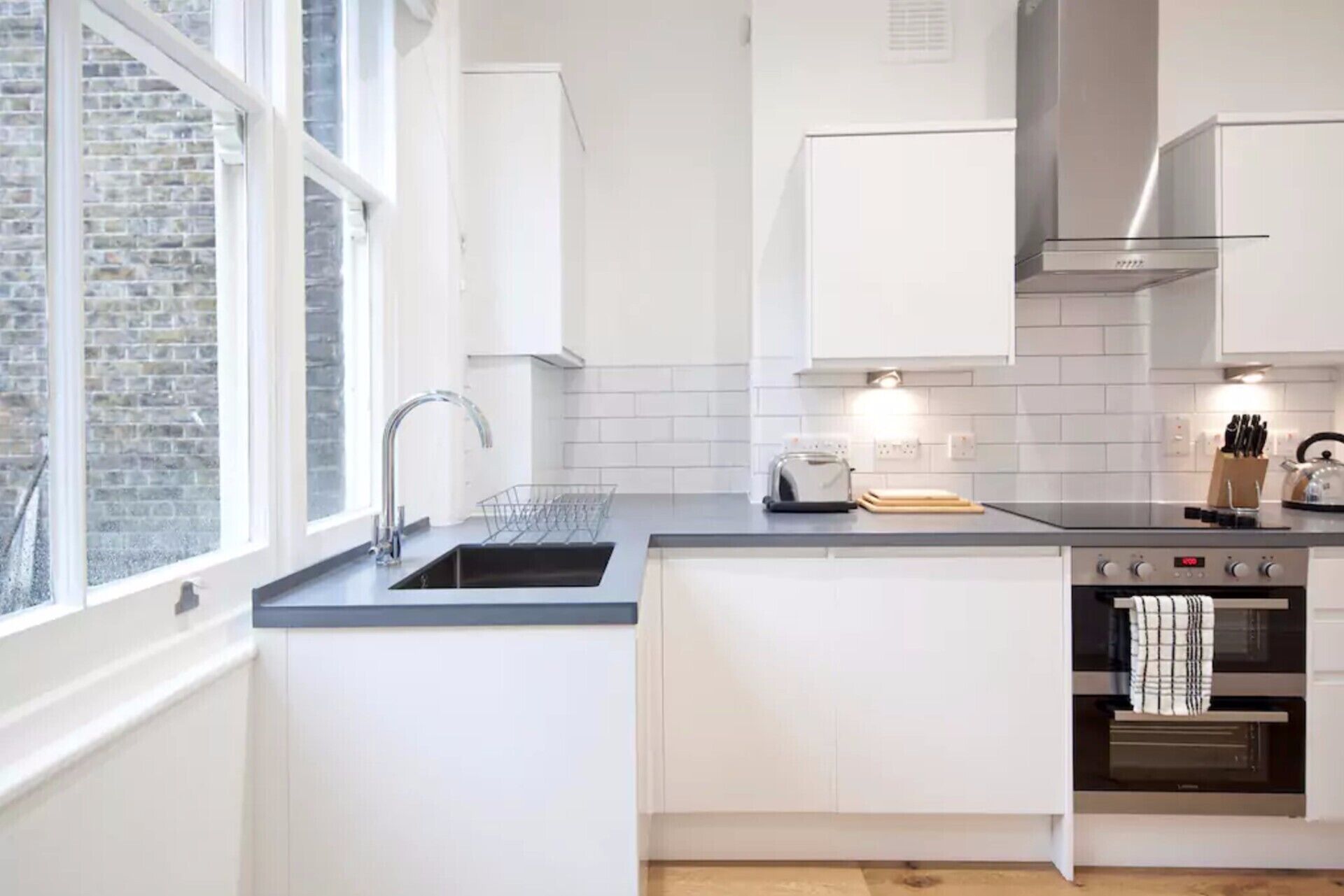 A modern kitchen with white cabinets, a stainless steel range hood, a built-in oven, and a window providing natural light.