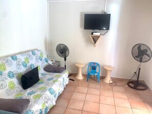 Une petite pièce comprenant un lit, un téléviseur fixé au mur, deux petites tables et deux ventilateurs.
