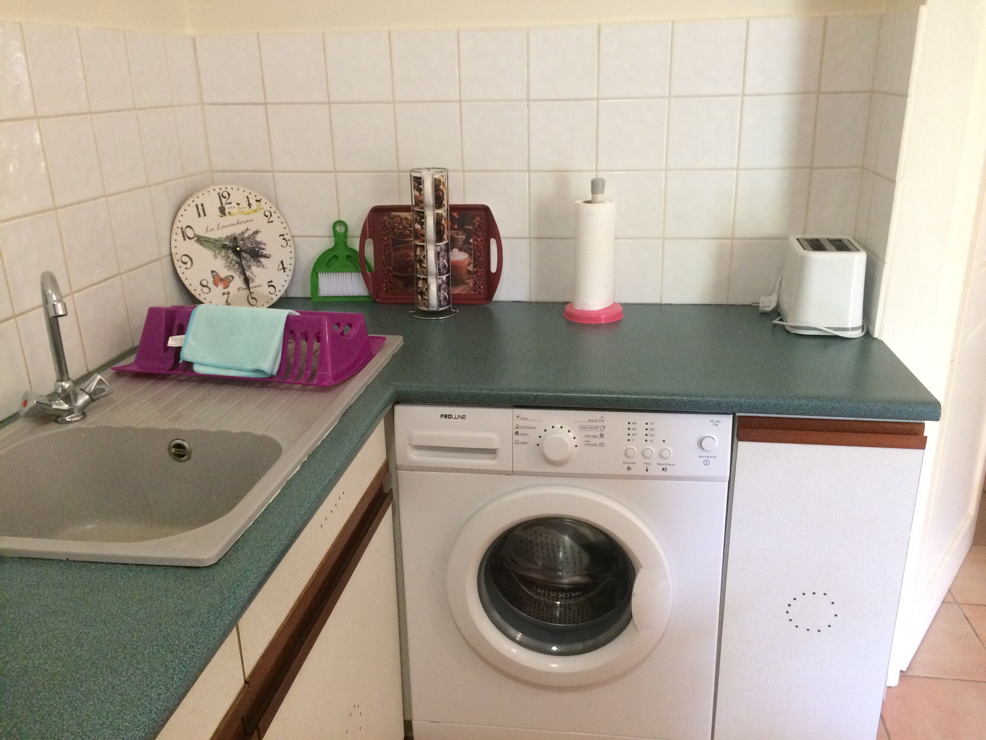 A compact kitchen with a sink, a washing machine, a toaster, and a clock on the wall.