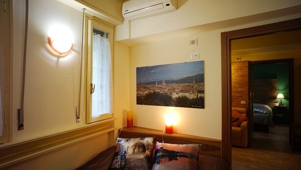 A hotel room with a bed, a window with curtains, a wall-mounted air conditioner, a framed cityscape picture, and a lit candle on a bedside table.