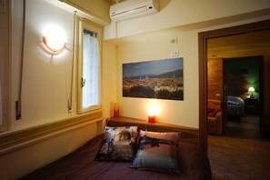 Habitación de hotel con cama, ventana con cortinas, aire acondicionado montado en la pared, una fotografía enmarcada de un paisaje urbano y una vela encendida sobre una mesita de noche.