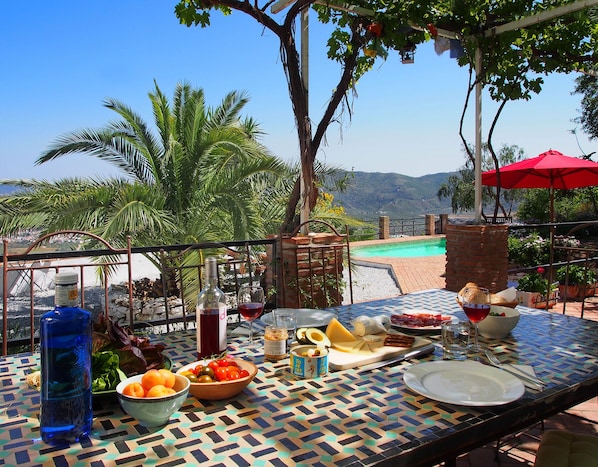 A table set outdoors with a view of a swimming pool and mountains, featuring a variety of fruits, cheeses, and a bottle of wine.