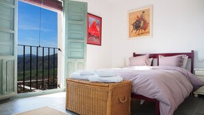 Une chambre à coucher avec un lit, un banc en osier, une vue sur l’extérieur et deux tableaux encadrés accrochés au mur.
