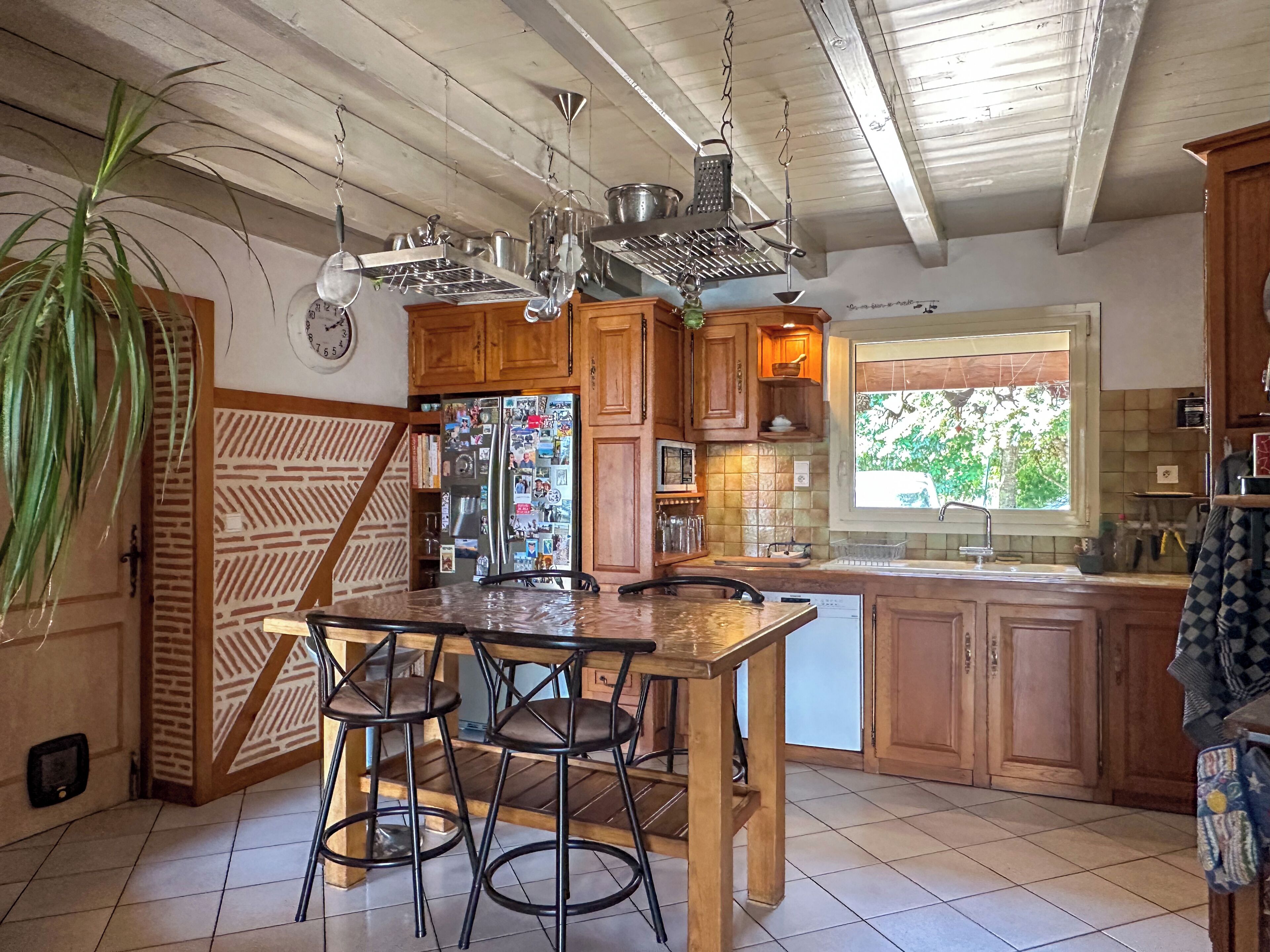 A kitchen with wooden cabinets, a stainless steel refrigerator, a wooden dining table with chairs, and a hanging light fixture.