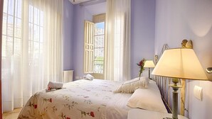 A bedroom with a bed, two bedside lamps, a window with curtains, and a view of buildings outside.