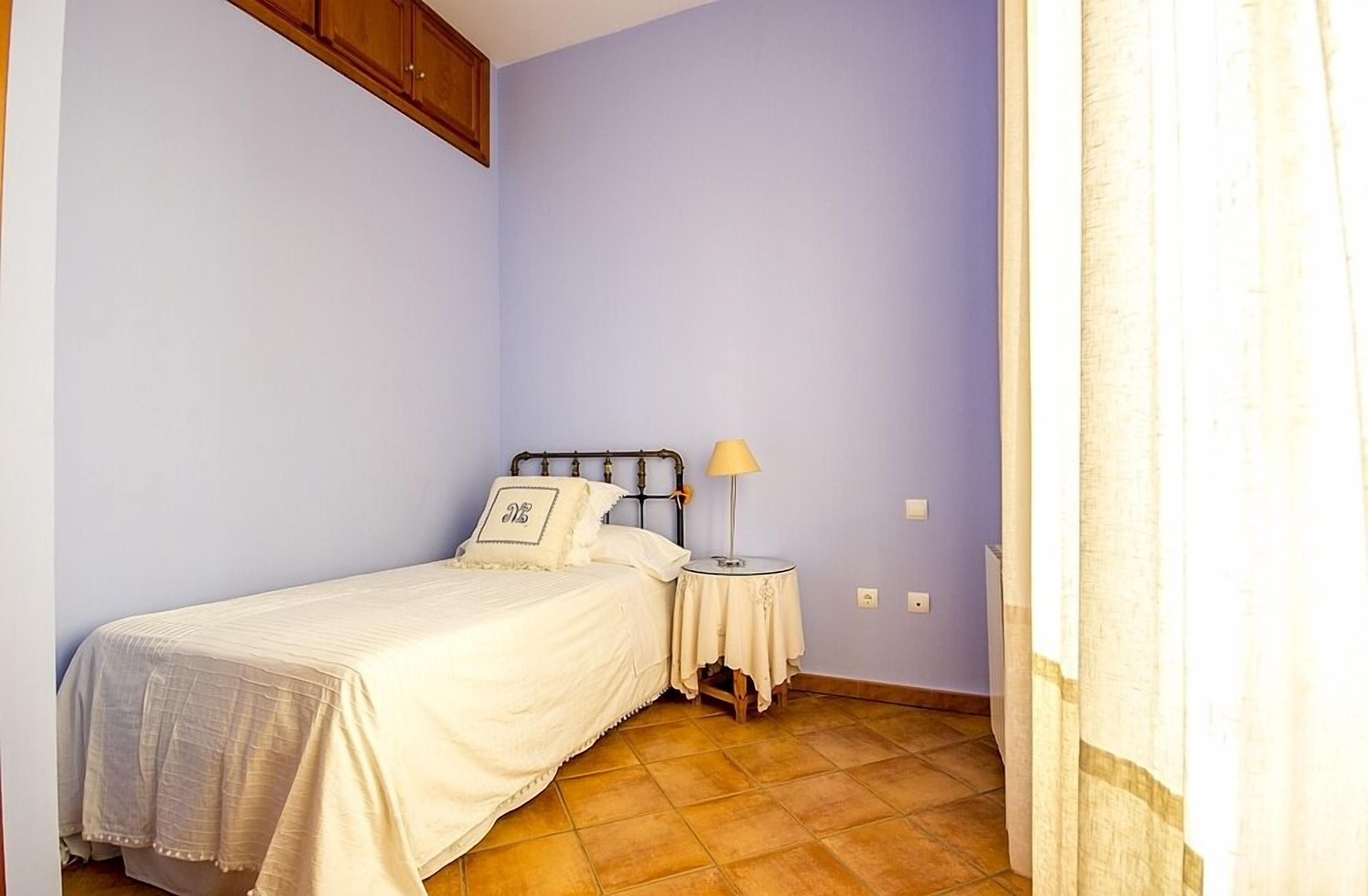 Una habitación pequeña con cama individual, cabecera de madera, mesita de noche con lámpara y pared lisa.