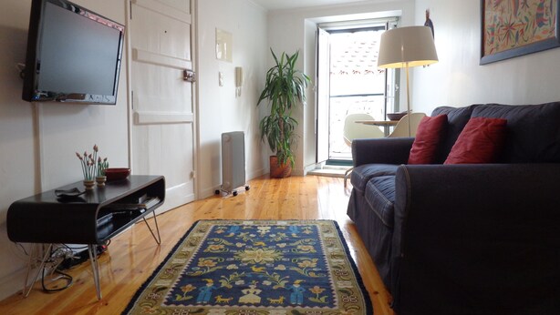 A living room with a dark-colored sofa, a flat-screen TV, a wooden floor, and a patterned rug. - Best in Alfama - Free pick up (Lisboa - Alfama)