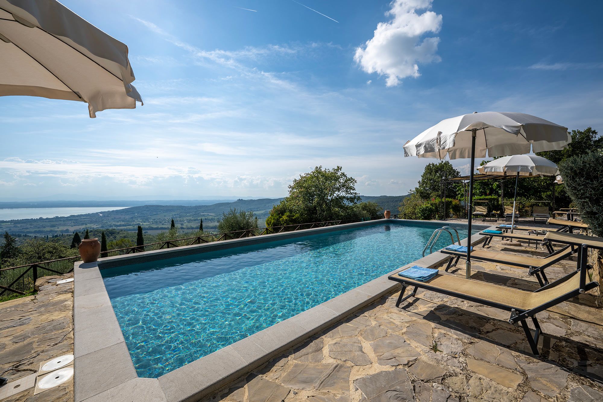 A swimming pool with clear blue water, surrounded by stone flooring, and overlooking a scenic landscape with trees and a distant body of water.