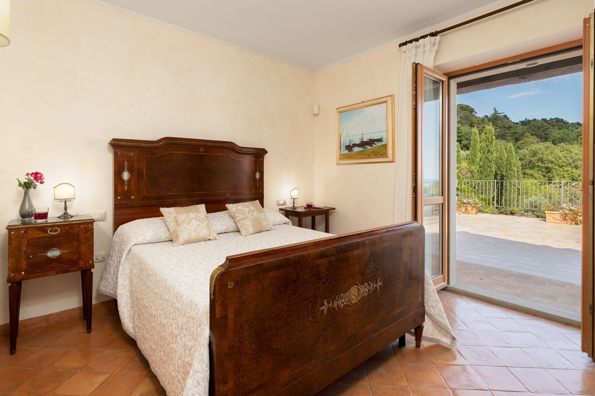 A bedroom with a wooden bed, a nightstand with a lamp, a bedside table with a vase and a glass, and a painting on the wall.