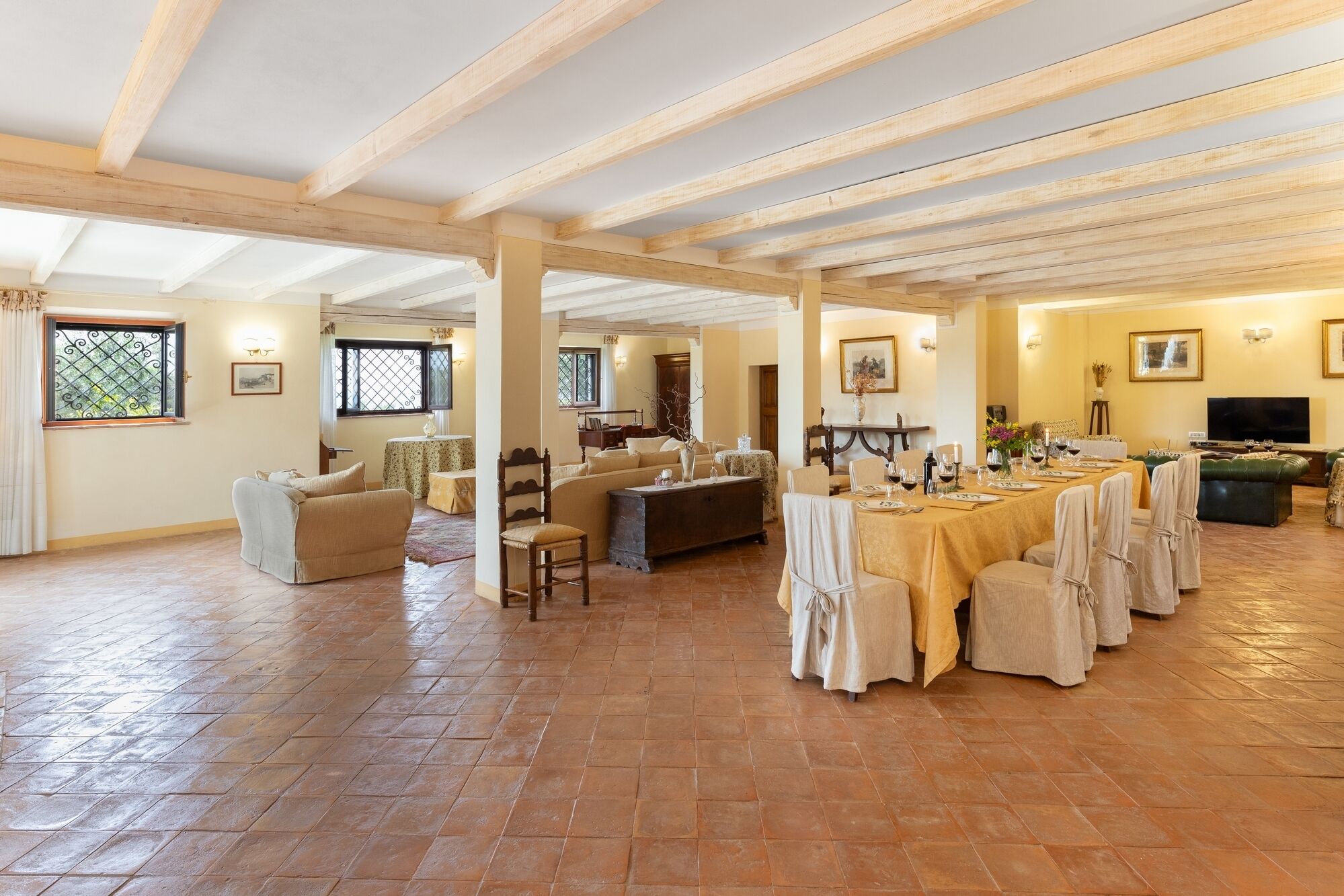 Ein geräumiges Zimmer mit hohen Decken und freiliegenden Holzbalken, ein langer Esstisch, gedeckt für eine Mahlzeit, und eine gemütliche Sitzecke mit Sofas und Sesseln.