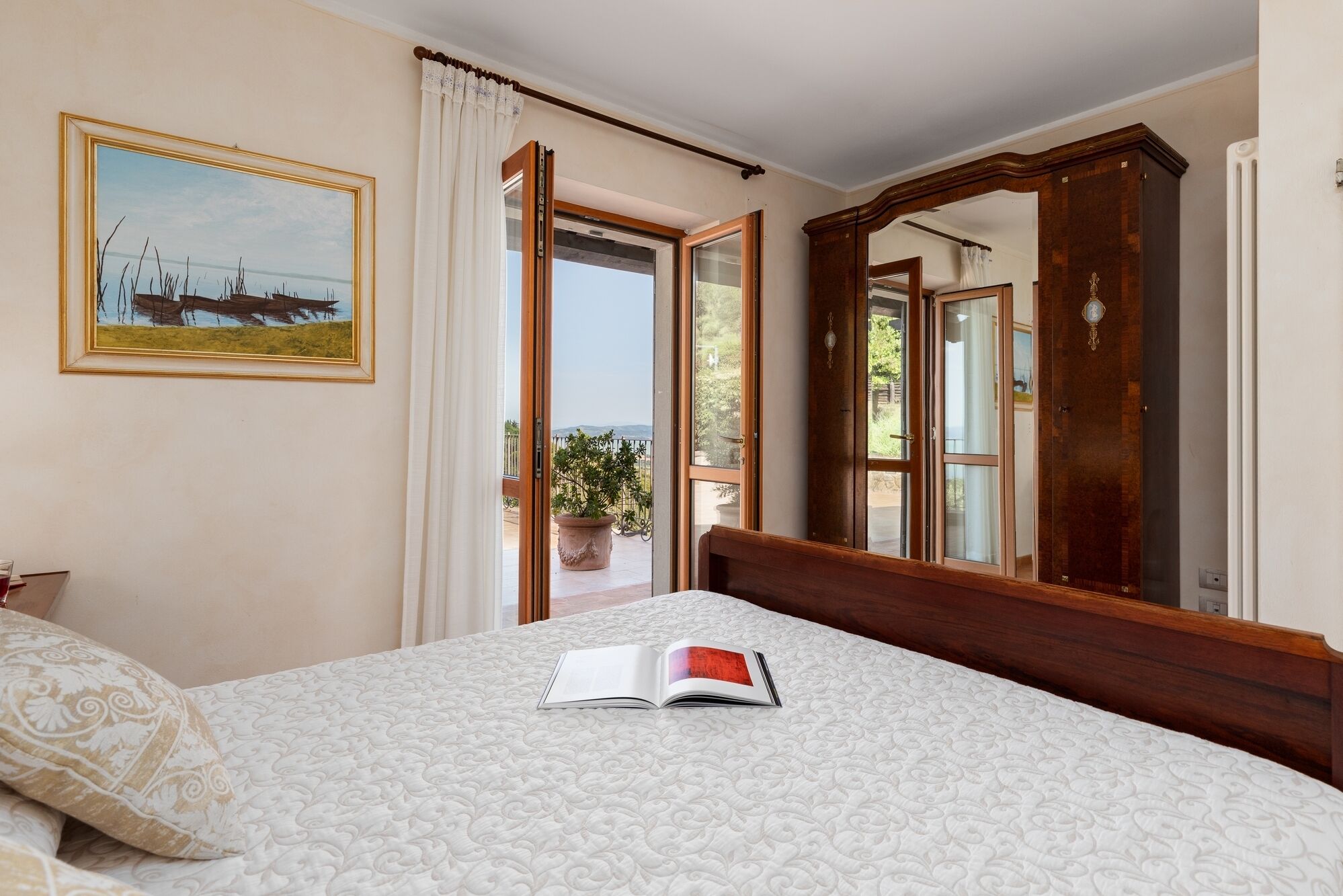 A bedroom with a bed, a large mirror, a painting on the wall, and an open door leading to a balcony with potted plants.