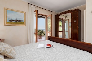 Een slaapkamer met een bed, een grote spiegel, een schilderij aan de muur en een open deur die leidt naar een balkon met potplanten.