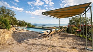 A stone patio with a pool, lounge chairs, and a covered area with a view of the sea.