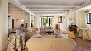 A spacious living room with a high ceiling, wooden beams, and a large window offering a view of greenery. There are multiple seating areas with floral-patterned tablecloths and a sofa.