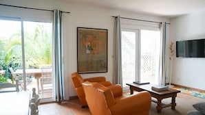 Ein Wohnzimmer mit einer Couch, einem Couchtisch und einem an der Wand montierten Fernseher. An der Wand hängt eine Karte, und eine Glastür führt in einen Außenbereich.