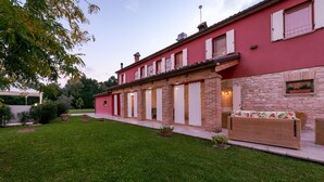 A red building with stone accents, a covered patio, and a grassy lawn. - Cascina Marianna 12+4, Emma Villas (Fossombrone)