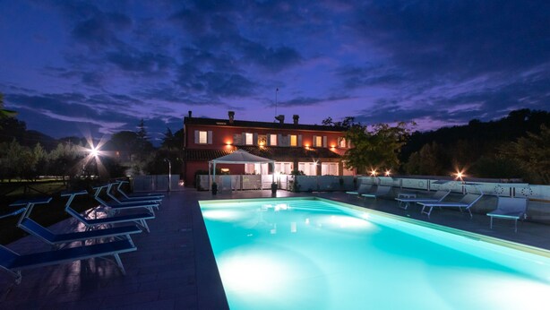 A swimming pool with lounge chairs, a covered seating area, and a building with multiple windows and a flag. - Cascina Marianna 12+4, Emma Villas (Fossombrone)