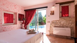 A bedroom with a bed, a stone fireplace, a window with red curtains, and a view of greenery outside. - Cascina Marianna 12+4, Emma Villas (Fossombrone)