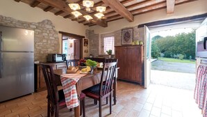 A rustic kitchen with stone walls, wooden beams, a dining table with chairs, and a view of the outdoors. - Cascina Marianna 12+4, Emma Villas (Fossombrone)