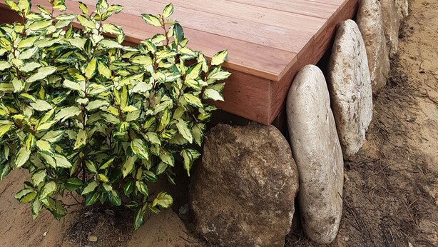 Une terrasse en bois avec un jardin intégré et des rochers, une table avec des chaises, et une clôture en bois en arrière-plan.