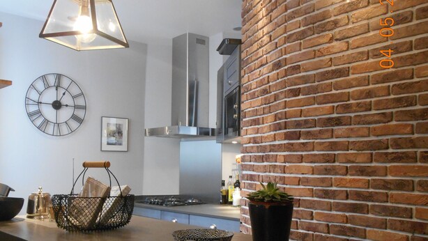 A modern kitchen with a brick wall, a hanging light fixture, a clock, and a dining area with chairs and a table.