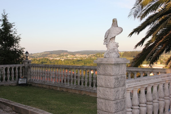 Una balaustrada de piedra con una estatua, con vistas a un paisaje pintoresco con árboles y una vista distante de edificios.