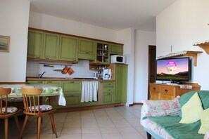A compact living space with a kitchenette, a dining area with two wooden chairs, a sofa with floral upholstery, and a television displaying a dessert advertisement.