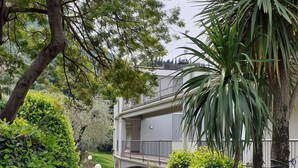 A pathway leading to a modern building with a balcony, surrounded by lush greenery and red flowers.