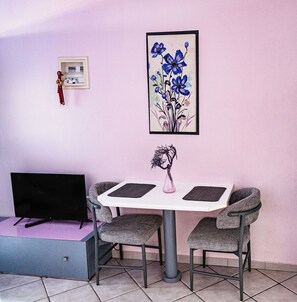 A dining area with a table, chairs, a vase with a plant, a framed artwork, and a television on a stand.