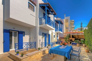 Un bâtiment traditionnel de couleur blanche, agrémenté de volets bleus et de balcons, une cour pavée de pierres et une table dressée pour le repas.