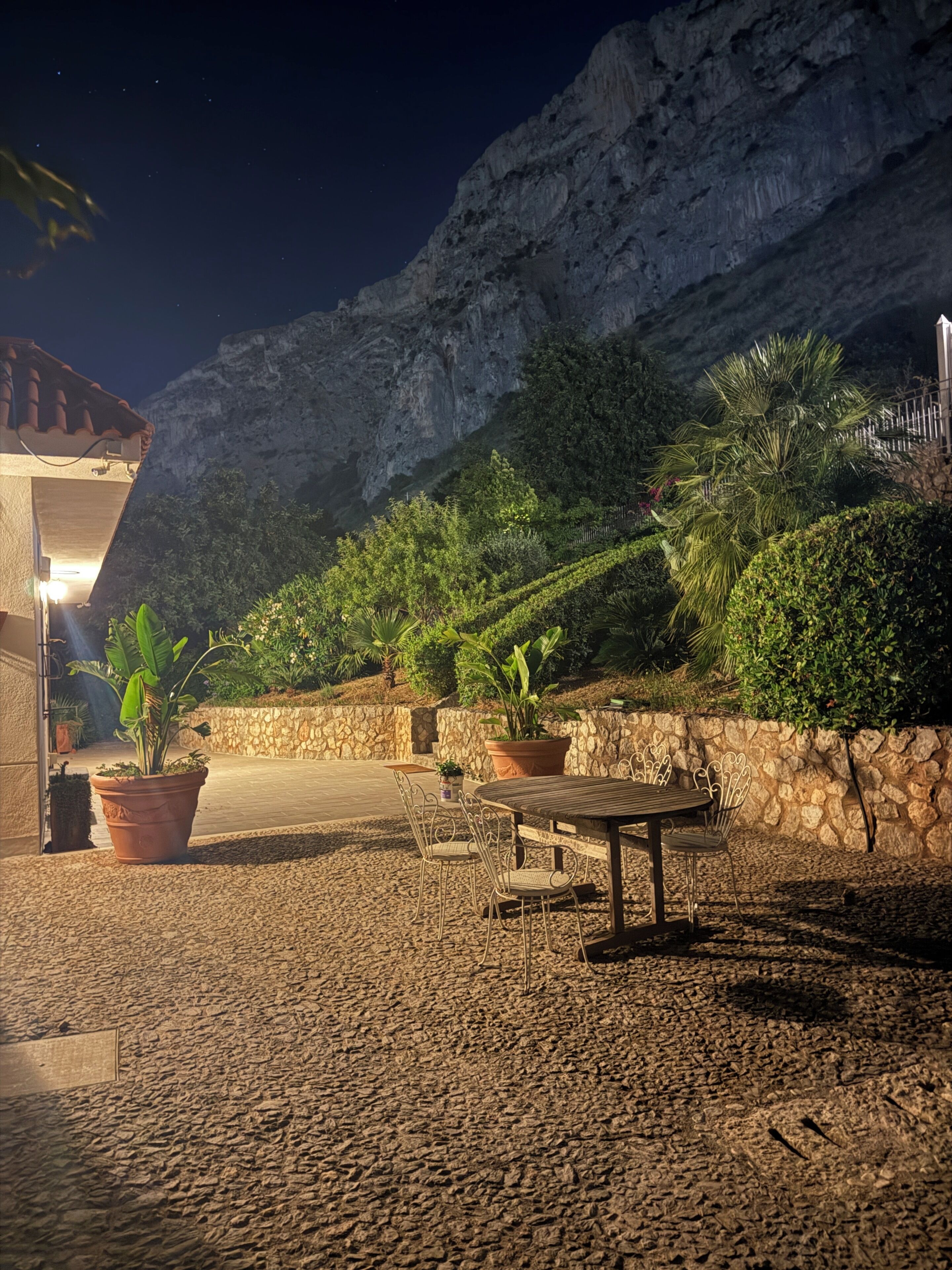 Un'area patio all'aperto con pavimento in pietra, piante in vaso, un tavolo in legno con sedie in metallo e un muro in pietra.