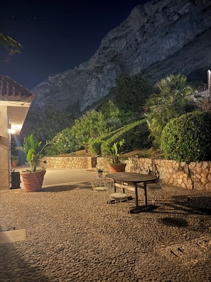 An outdoor patio area with a stone floor, potted plants, a wooden table with metal chairs, and a stone wall.