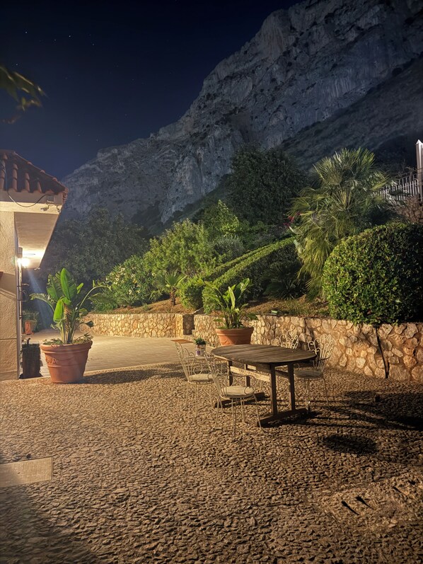 An outdoor patio area with a stone floor, potted plants, a wooden table with metal chairs, and a stone wall.