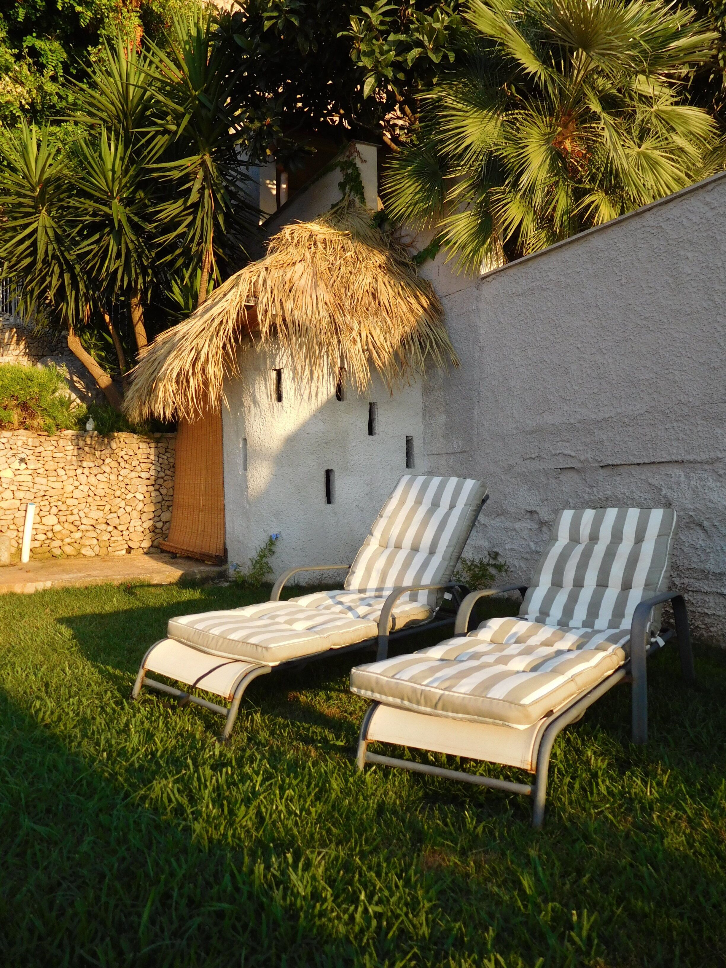 Dos sillones con cojines a rayas, un techo de paja y un muro de piedra en un jardín.