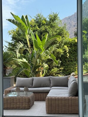 A modern outdoor seating area with a grey sofa and wicker furniture, surrounded by lush greenery and a mountain backdrop. - Villa between sea and mountain with private pool and sea view with private pool  (Capaci)