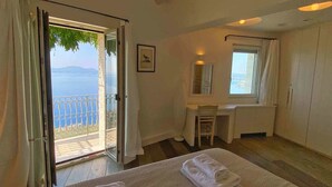 A hotel room with a bed, a desk, a chair, a window with a view of the sea, and a balcony with a railing. - Paleopetres Marnie - Private Pool - Sea Views - Beaches - North East Coast - (Nisaki)