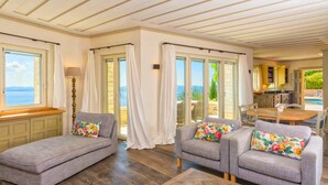 A spacious living area with a view of the sea, a wooden floor, and a kitchen visible through an open door. - Paleopetres Marnie - Private Pool - Sea Views - Beaches - North East Coast - (Nisaki)