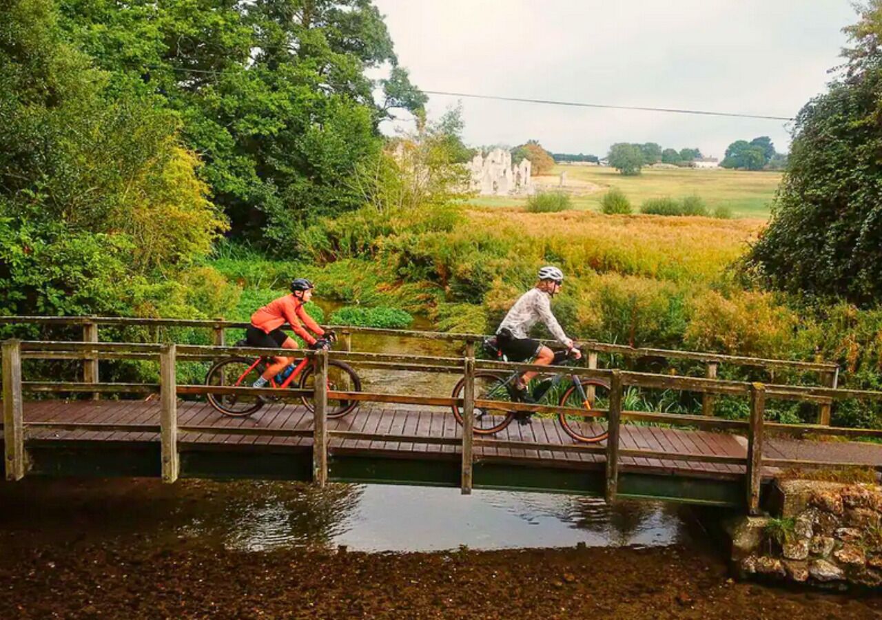 Two cyclists on a wooden bridge over a water body with greenery and a distant building in the background.