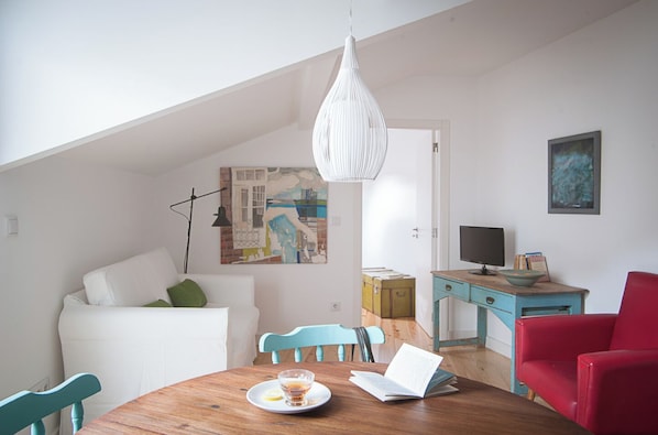 Um espaço de estar acolhedor com um sofá branco, uma mesa de jantar de madeira, uma cadeira vermelha, uma cadeira azul, uma escrivaninha verde-água e uma luminária pendente branca.
